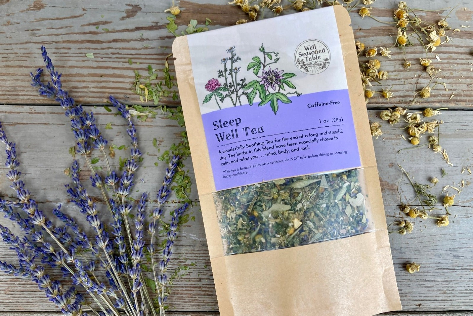  A packet of Sleep Well Tea from Well Seasoned Table on a wooden background with dried lavender and chamomile flowers around it.