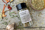 A glass shaker jar of Spice of Life (everything) Seasoning from Well Seasoned Table on a wooden background with a bowl of seasoning, dried peppers, garlic, and herbs.
