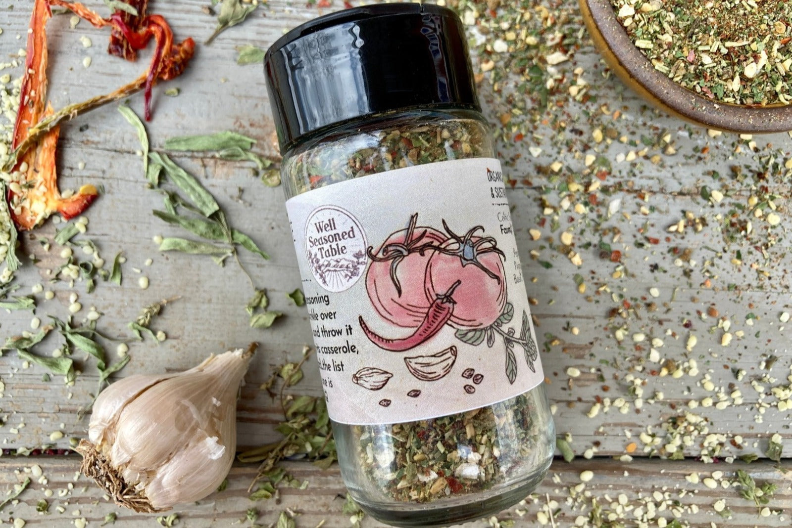 The side of a glass shaker jar of Spice of Life (everything) Seasoning from Well Seasoned Table on a wooden background with a bowl of seasoning, dried peppers, garlic, and herbs.