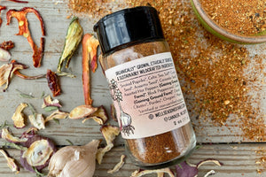 The back of a glass shaker jar of Taco Seasoning from Well Seasoned Table on a wooden background with dried peppers, garlic, and seasoning around it.