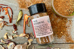 A glass shaker jar of Taco Seasoning from Well Seasoned Table on a wooden background with dried peppers, garlic, and seasoning around it.