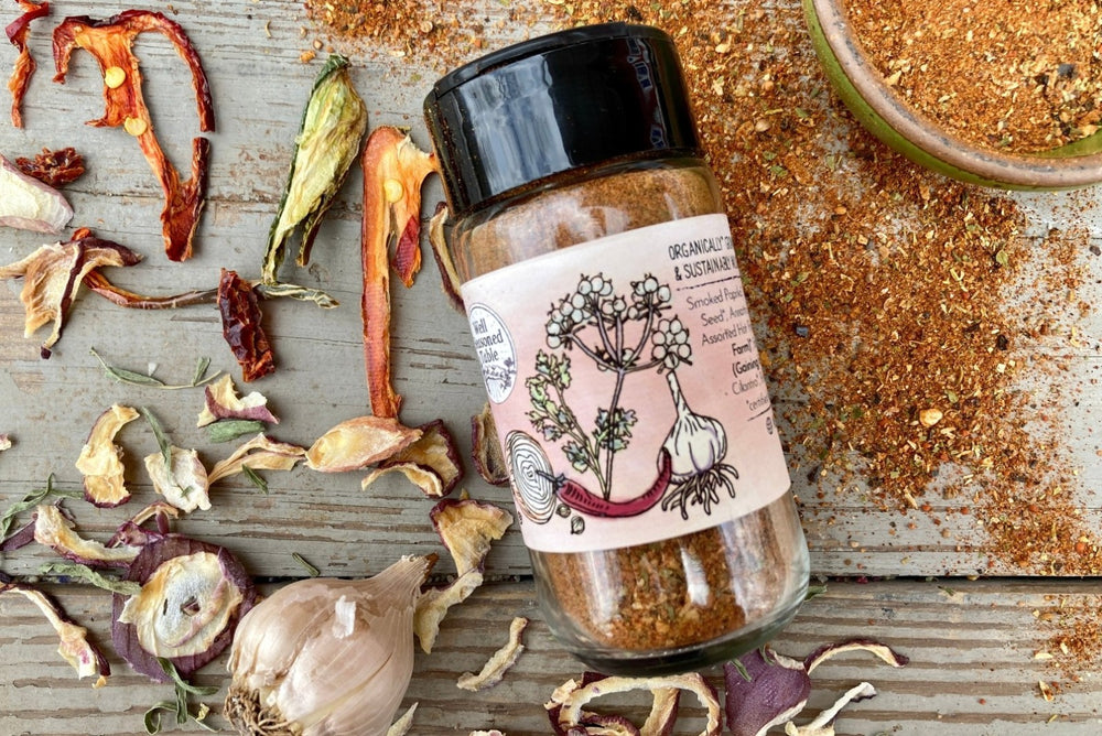 The side of a glass shaker jar of Taco Seasoning from Well Seasoned Table on a wooden background with dried peppers, garlic, and seasoning around it.