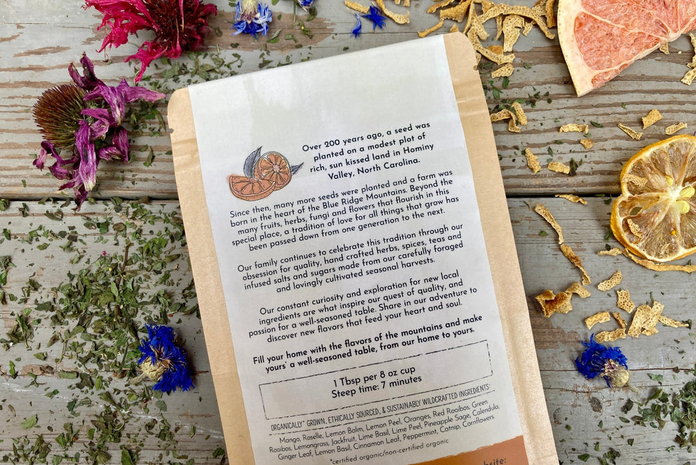 The back of a packet of Tropical Tango Tea from Well Seasoned Table on a wooden background surrounded by dried herbs, flowers, and fruits.