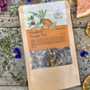 A packet of Tropical Tango Tea from Well Seasoned Table on a wooden background surrounded by dried herbs, flowers, and fruits.