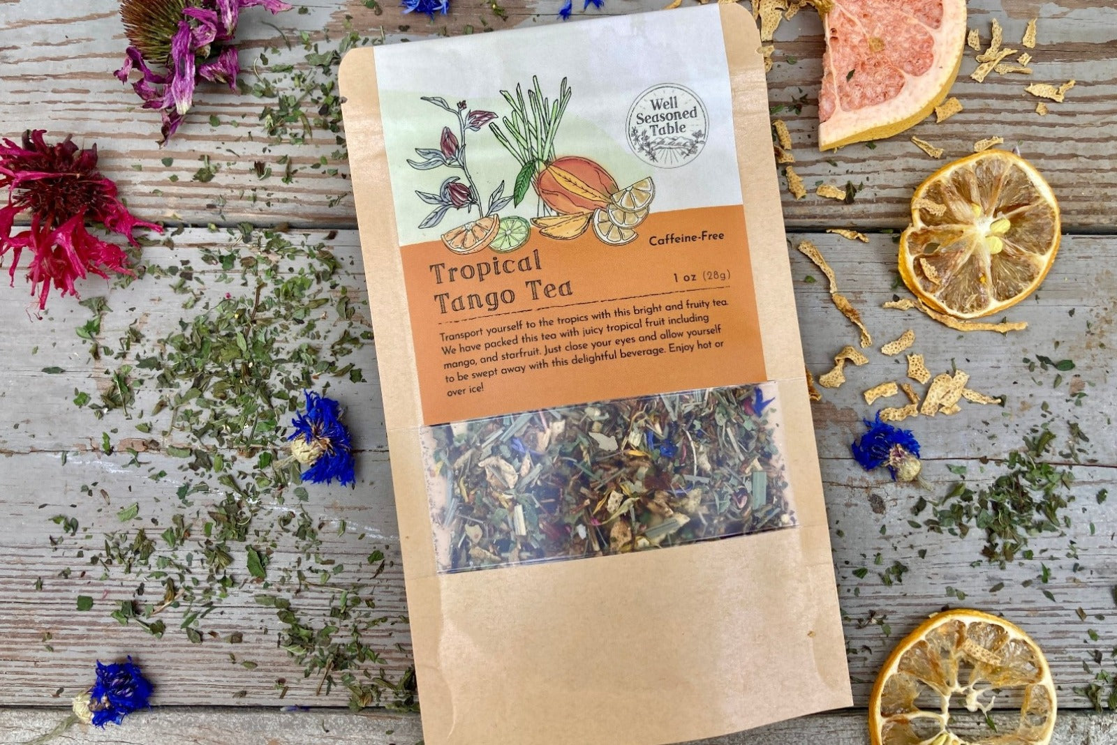 A packet of Tropical Tango Tea from Well Seasoned Table on a wooden background surrounded by dried herbs, flowers, and fruits.