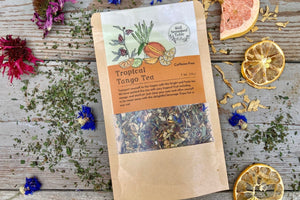 A packet of Tropical Tango Tea from Well Seasoned Table on a wooden background surrounded by dried herbs, flowers, and fruits.