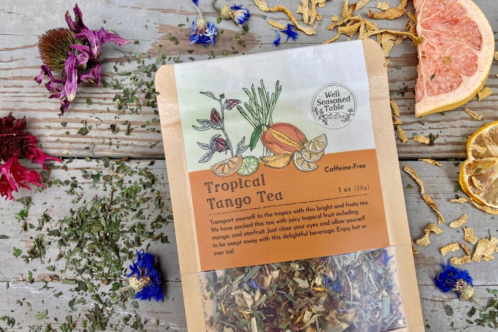 Up close of a packet of Tropical Tango Tea from Well Seasoned Table on a wooden background surrounded by dried herbs, flowers, and fruits.