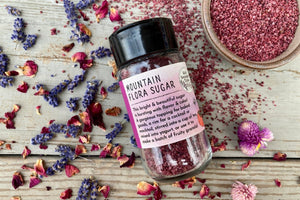 A glass shaker jar of Mountain Flora Sugar from Well Seasoned Table on a wooden background with dried lavender and roses, and a ceramic bowl of bright rose colored sugar.