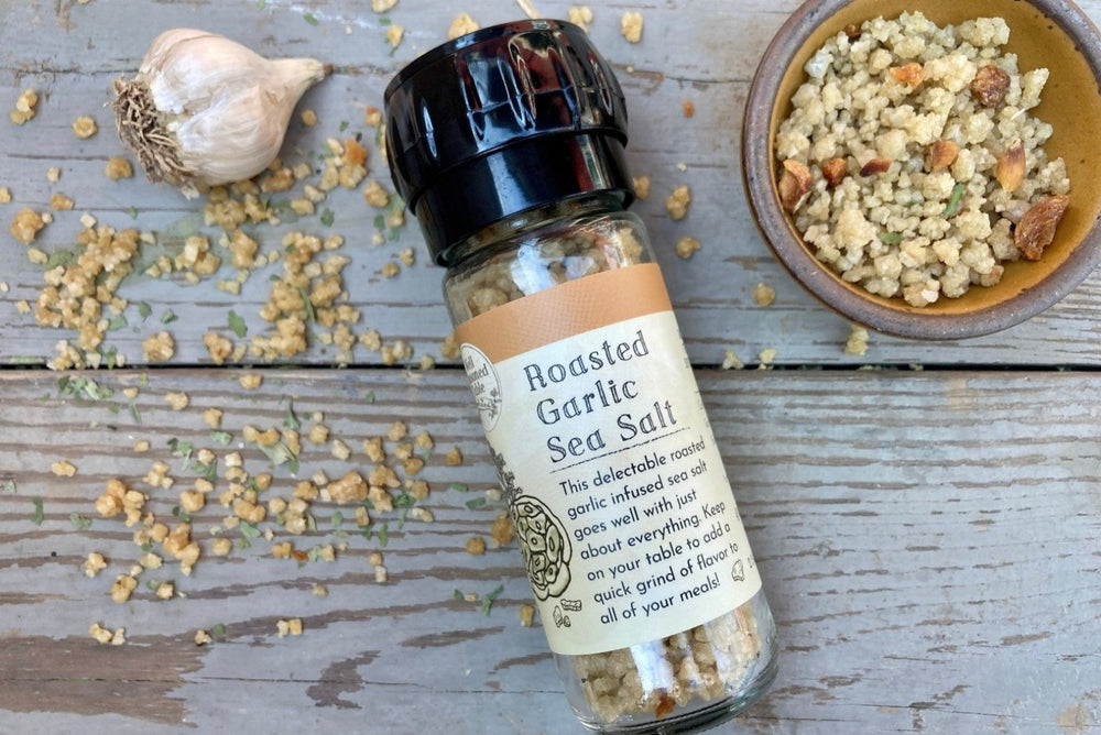 A glass grinder jar of Roasted Garlic Sea Salt on a wooden background with a bowl of the infused sea salt from Well Seasoned Table.