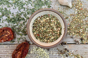 A ceramic bowl of Spice of Life (everything) Seasoning from Well Seasoned Table on a wooden background with dried garlic, tomatoes, and herbs around it.