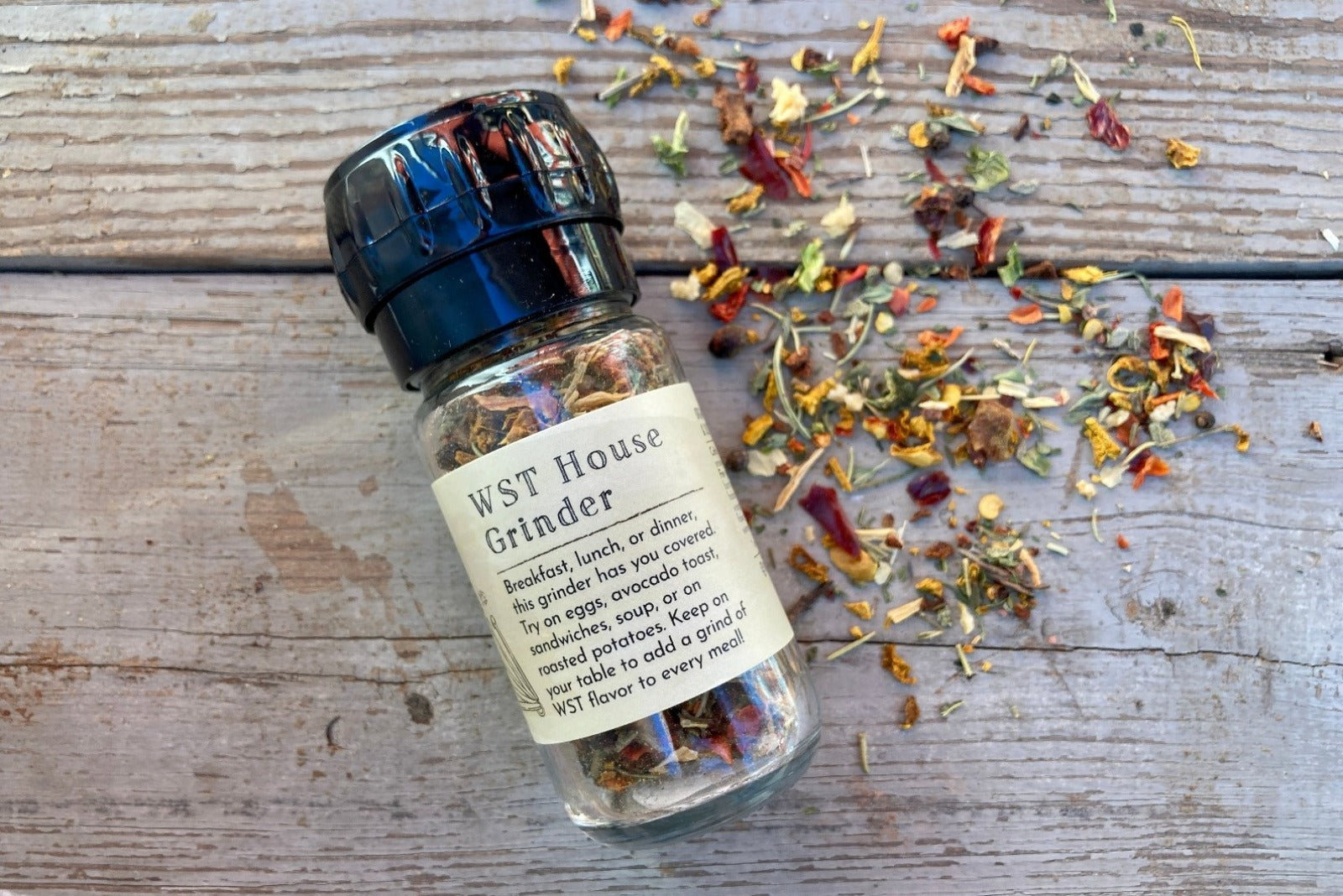 A glass grinder jar of WST House Grinder from Well Seasoned Table on a wooden background with seasonings sprinkled around it.