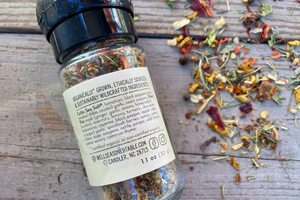 A glass grinder jar of WST House Grinder from Well Seasoned Table on a wooden background with seasonings sprinkled around it.