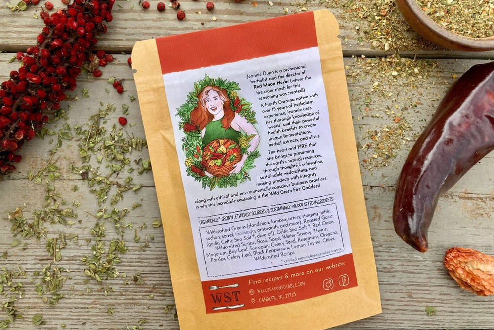 The back of a packet of Wild Green Fire Goddess Seasoning on a wooden background with dried sumac, peppers, and herbs.