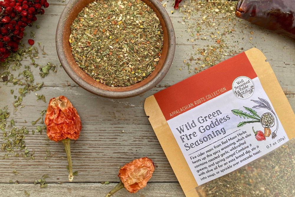 A packet and a bowl of Wild Green Fire Goddess Seasoning on a wooden background with dried sumac, peppers, and herbs.