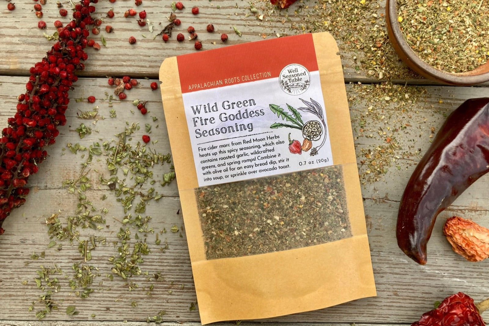 A packet of Wild Green Fire Goddess Seasoning on a wooden background with dried sumac, peppers, and herbs.