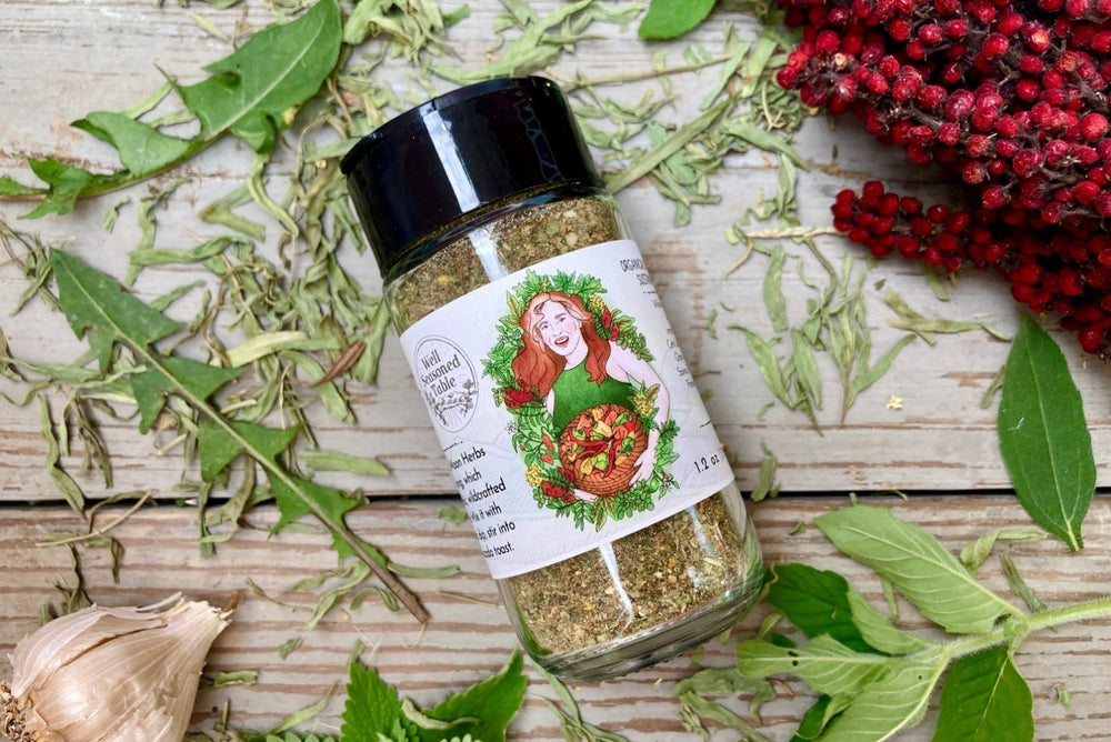 The side of a glass shaker jar of Wild Green Fire Goddess from Well Seasoned Table on a wooden background with sumac, wild greens and garlic around it.