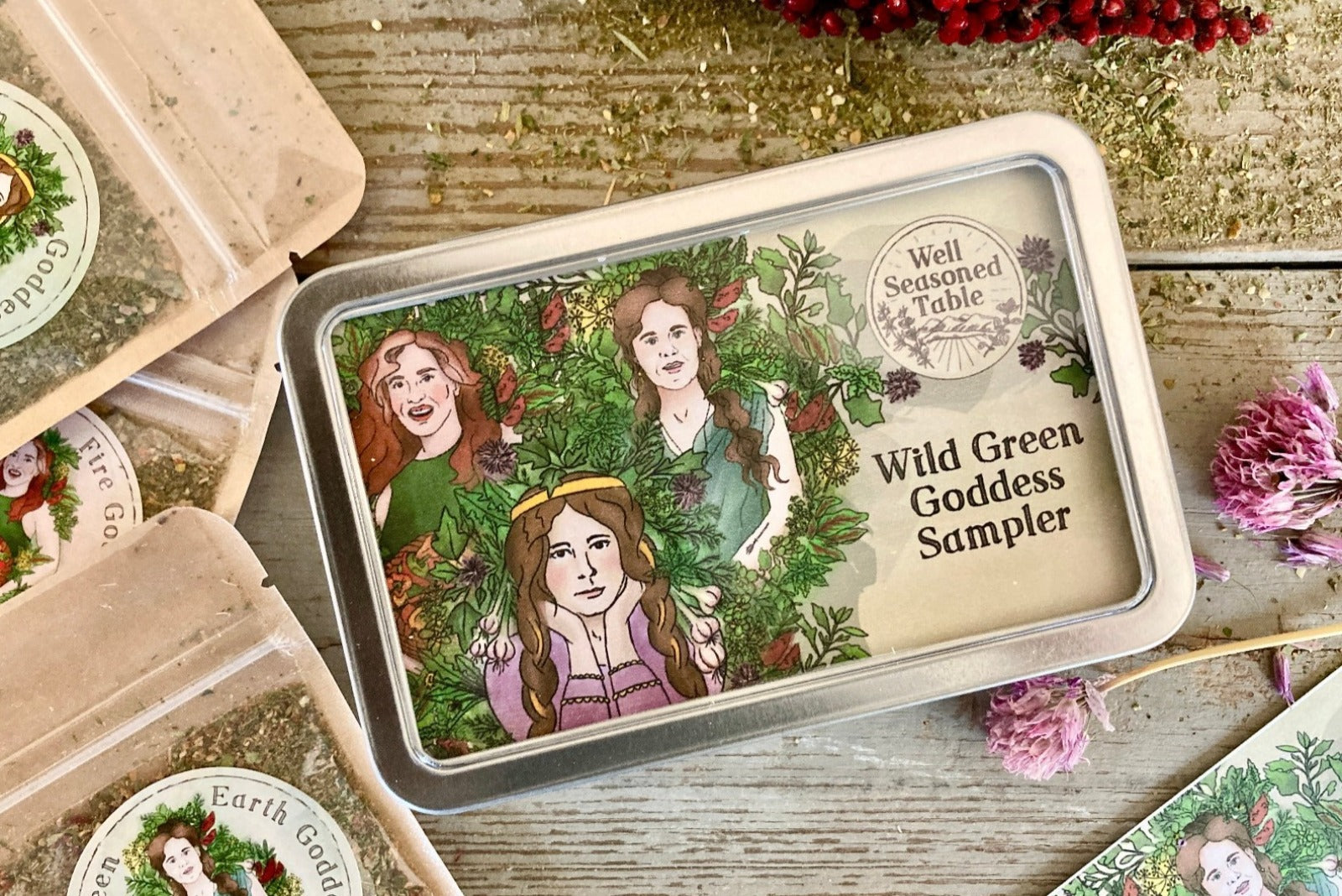 A sampler tin of Wild Green Goddess seasonings from Well Seasoned Table with packets of seasonings and dried spices around it.