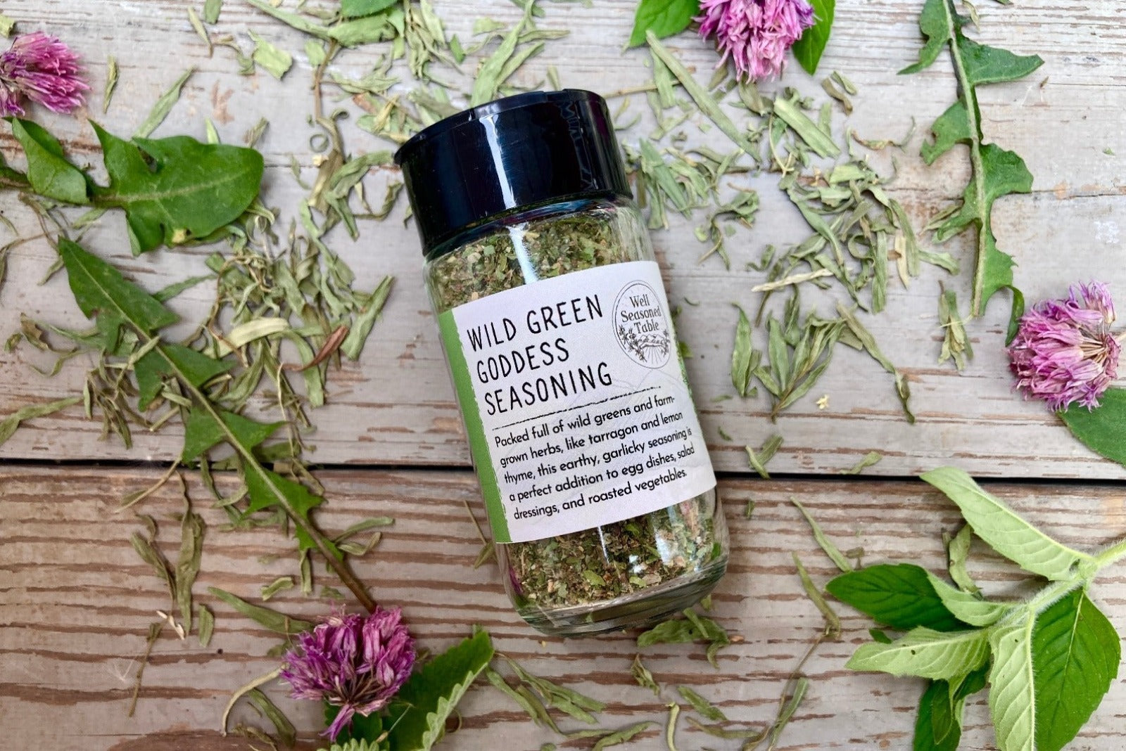 A glass shaker jar of Wild Green Goddess Seasoning from Well Seasoned Table on a wooden background with wild greens and chive blossoms around it.