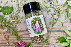 The side of a glass shaker jar of Wild Green Goddess Seasoning on a wooden background with wild greens and chive blossoms around it.