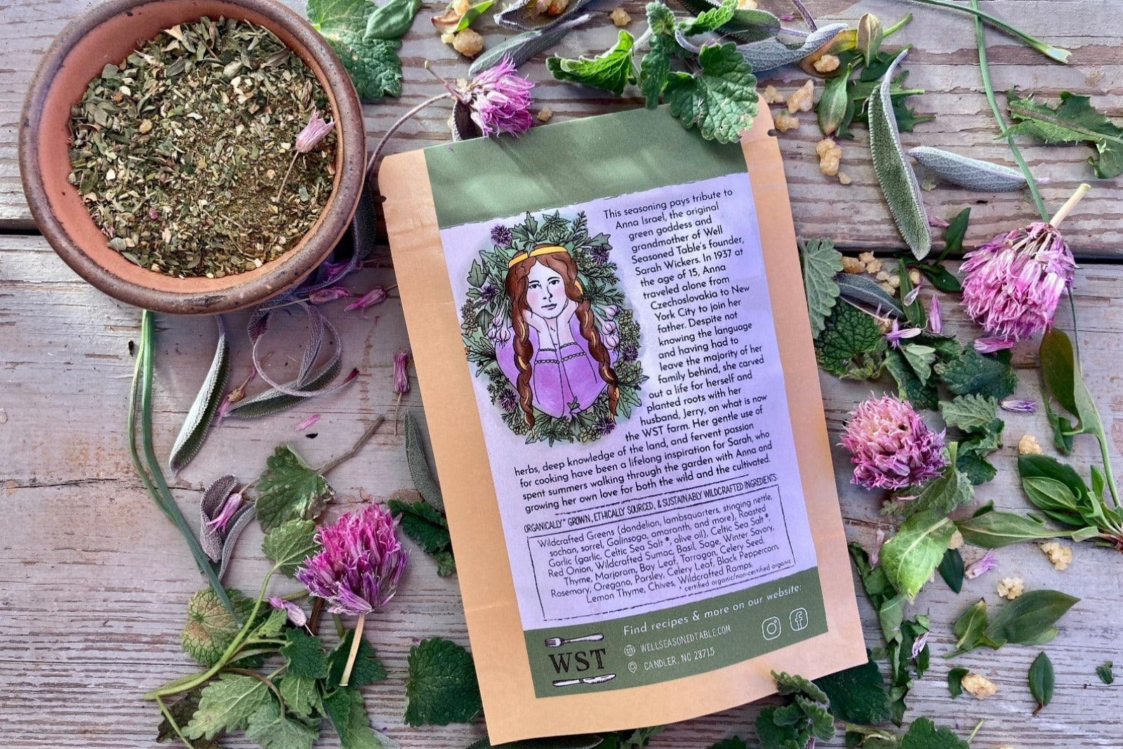 The back of a packet of Wild Green Goddess Seasoning from Well Seasoned Table on a wooden background with wild greens and chive blossoms around it.