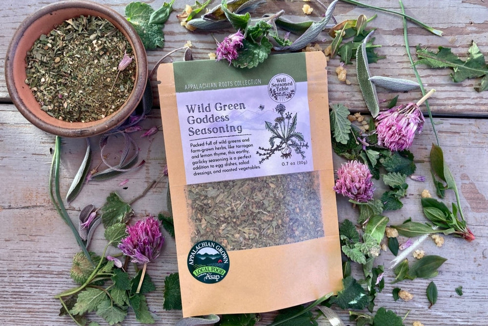 A packet of Wild Green Goddess Seasoning from Well Seasoned Table on a wooden background with wild greens and chive blossoms around it.