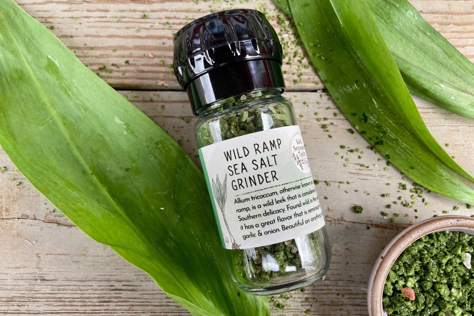 A glass jar of Wild Ramp Sea Salt in a grinder jar from Well Seasoned Table on a wooden background with a small bowl of Wild Ramp Sea Salt.