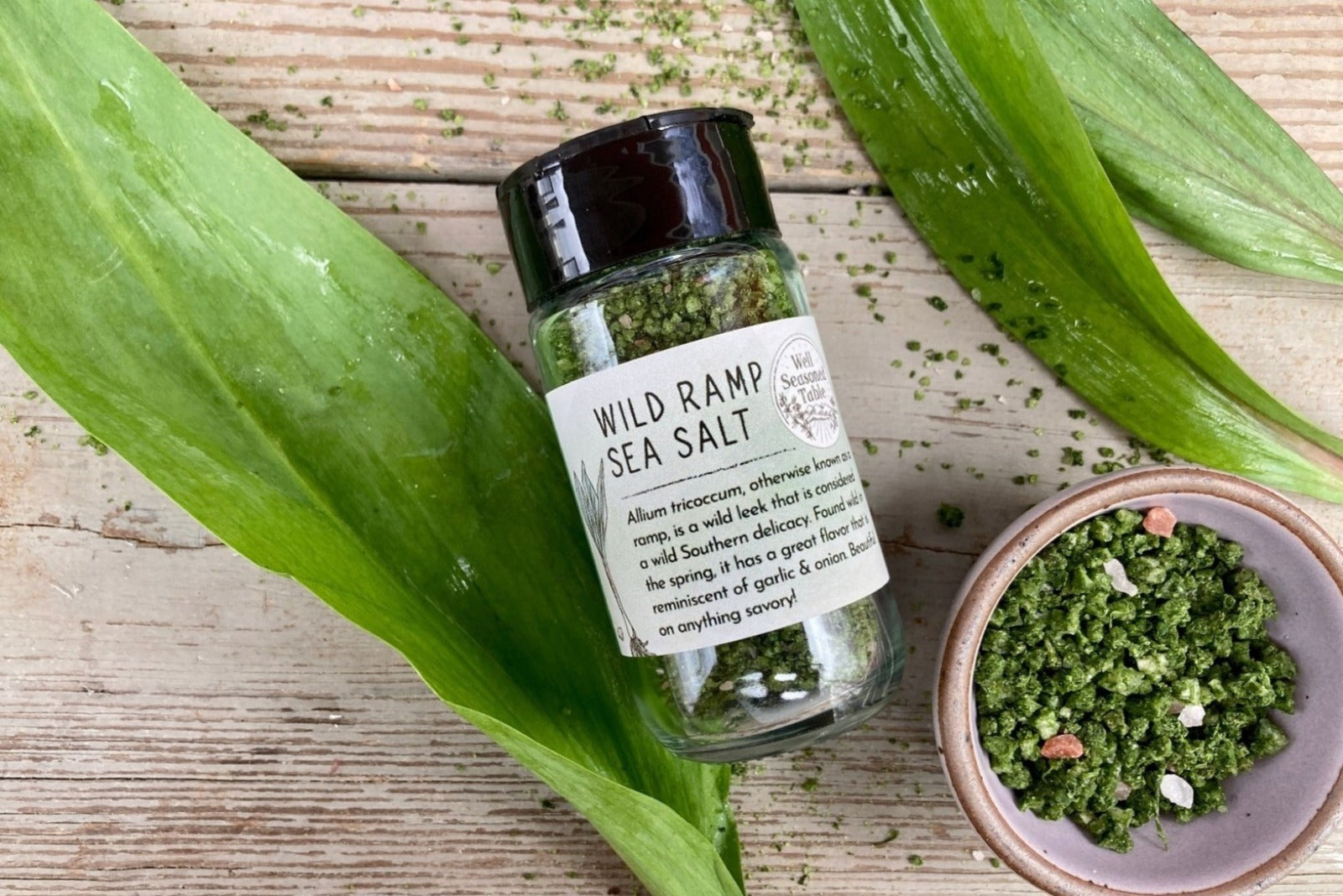 A glass shaker jar of Wild Ramp Sea Salt from Well Seasoned Table on a wooden background with a green ceramic mini bowl of bright green infused sea salt.