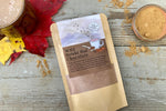A packet of Wild Reishi Hot Chocolate from Well Seasoned Table on a wooden background with a bowl of cocoa powder.